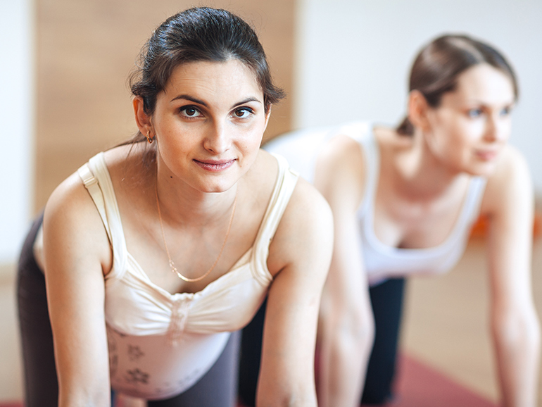 Two pregnant women during a pilates class on all fours stretching, one woman is smiling at the camera with the other in the background not quite in focus