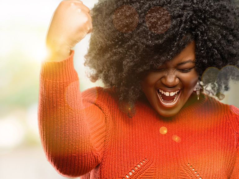 A woman is outside with the sunlight behind her and has her arm raised with her fist clenched in a celebratory position. Her mouth is open and her face clenched as if shouting something celebratory.