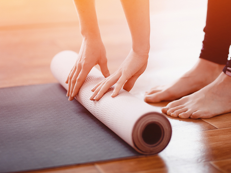 A yoga or exercise mat is being rolled up on a wooden floor by a woman whose hands and feet are in the shot. She appears to be wearing leggings.