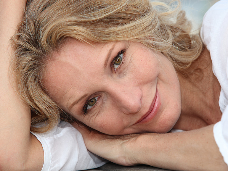 An older woman is shown smiling while lying down with her head supported by her arms in a white top, representing a menopausal woman.