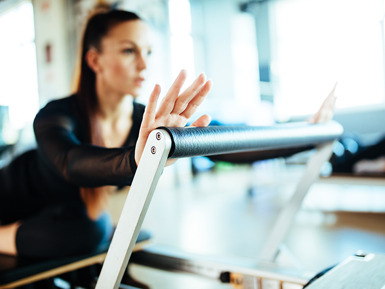 A young woman is stretching using equipment in a studio, slightly out of focus wearing black exercise clothes.