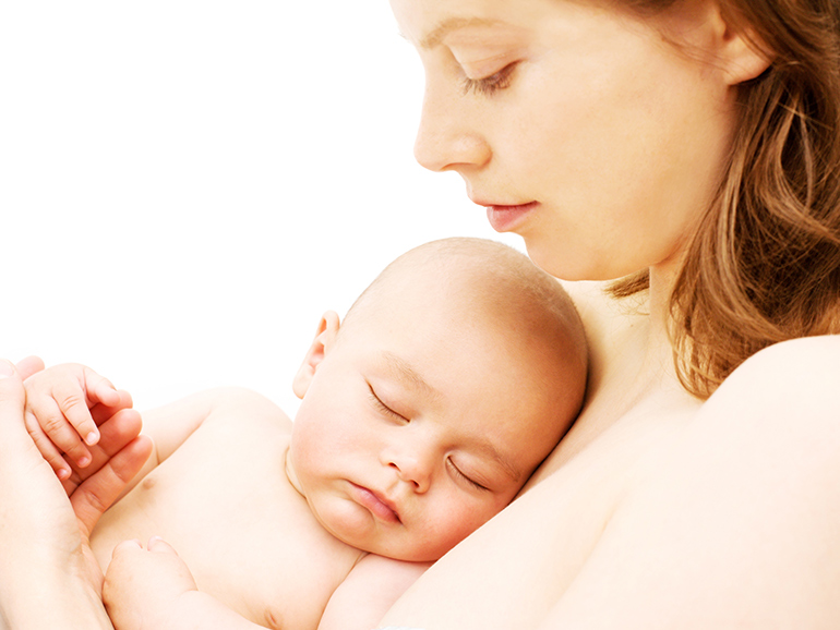 A shot of a woman's head and shoulders holding her sleeping baby who is resting on her chest. The mother is looking down at her baby.