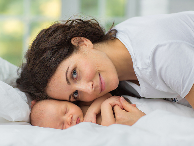 A woman with brown hair in a white top is lying down smiling next to her sleeping newborn baby.