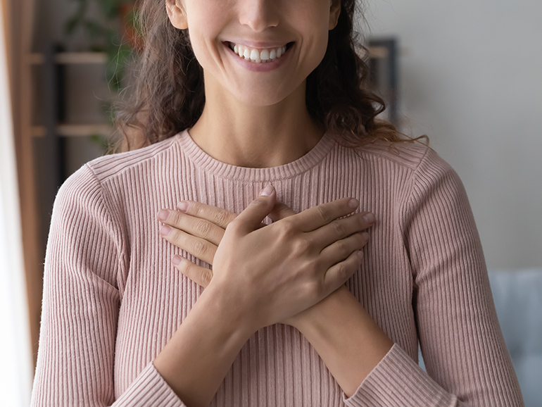 A woman's upper body and partial head are shown in frame. The woman is smiling and her hands are held over her heart in a caring gesture.