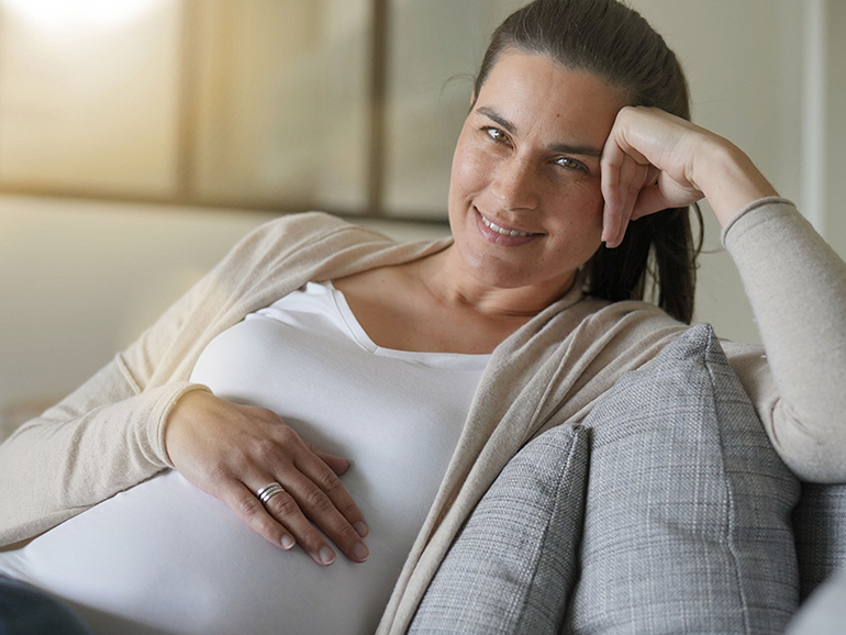 A pregnant woman is sitting on a sofa looking comfortable in a white top and cream cardigan. She is resting her head on her hand and smiling at the camera.